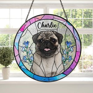 May include: A stained-glass window hanging features a pug dog portrait with the name Charlie. The round design incorporates pink, purple, and blue hues, accented with blue flowers. The pug is centrally positioned, detailed with textured glass, against a textured background. The piece is framed with a black metal hanging ring.