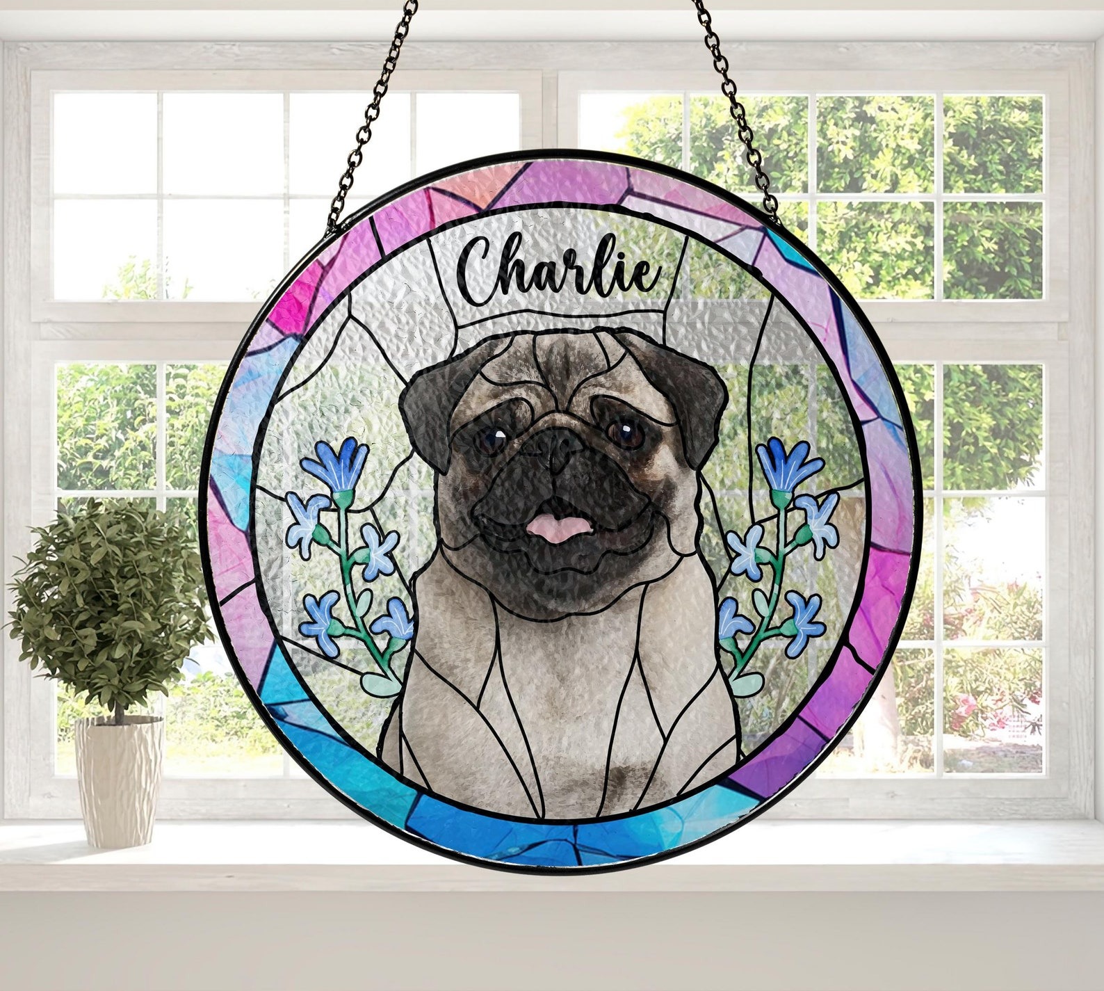 7 May include: A stained-glass window hanging features a pug dog portrait with the name Charlie. The round design incorporates pink, purple, and blue hues, accented with blue flowers. The pug is centrally positioned, detailed with textured glass, against a textured background. The piece is framed with a black metal hanging ring.